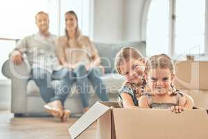 Two cute little girls kids playing with cardboard boxes at new apartment. Caucasian parents sitting with laptop on couch on the background. Relocation and moving day of big family