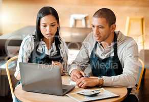 Two young mixed race employees wearing aprons sitting at a table in a coffee shop using a laptop and calculator at work. Young male and female hispanic colleagues brainstorming as a team because alone we can do so little, together we can achieve so much