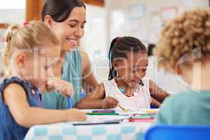 Teaching and kids looking happy while doing activity at kindergarten or pre-school. Smiling teacher sitting at table with toddlers while colouring with crayons
