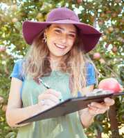 Portrait of female farm worker holding an apple while writing and making notes on a orchard farm during harvest season. Agronomist doing inspection and record keeping on the quality of produce