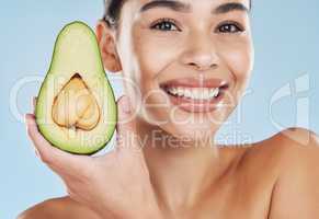 Beautiful young mixed race woman with an avocado isolated in studio against a blue background. Her skincare regime keeps her fresh. For glowing skin, eat healthy. Packed with vitamins and nutrients