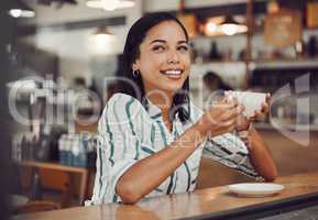 Happy young smiling mixed race woman in casual outfit drinking hot beverage while sitting alone at a table in cozy cafe. Beautiful female looking happy and thoughtful drinking coffee and enjoying her leisure time alone