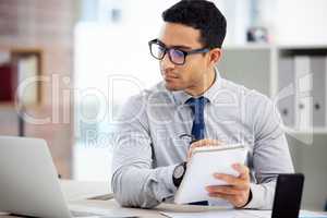 Focused mixed race businessman writing in a diary while working on a laptop alone in an office at work. One hispanic male businessperson making a list in a notebook while sitting at a desk at work