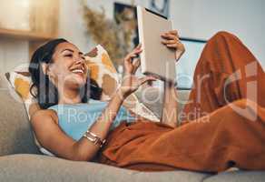 Smiling mixed race woman browsing internet on digital tablet at home. Happy hispanic lying down on living room sofa alone and using technology. Relaxed woman scrolling and searching online on weekend