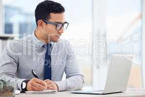 Focused mixed race businessman writing in a diary while working on a laptop alone in an office at work. One serious hispanic male businessperson making a list in a notebook while sitting at a desk at work