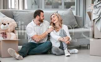 Young cheerful caucasian couple hugging and laughing while sitting on the floor of their new house in the lounge. Joyful husband smiling and giving his wife a hug after moving into a new apartment