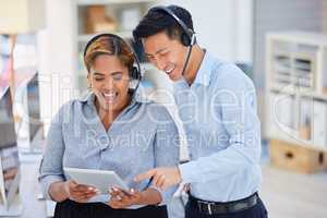 Two happy young diverse call centre telemarketing agents working on a digital tablet in an office. Friendly colleagues troubleshooting solution for customer service and sales support while operating helpdesk together