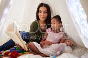 Spark your childs imagination through stories. a mother reading a book to her little daughter under a blanket fort at home.