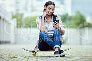 Checking her messages on the fly. Full length shot of an attractive young female skater checking her messages while sitting on her skateboard outside.