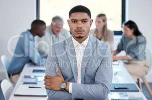 I am not afraid to take risks where needed. Portrait of a confident young businessman standing with his arms crossed in an office while his colleagues have a meeting in the background.