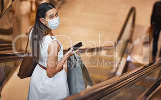 Portrait of a young mixed race woman wearing a medical face mask and holding a cellphone and shopping bags while on an escalator. Serious hispanic woman looking back over shoulder while carrying retail bags after buying in a mall during Covid-19 pandemic