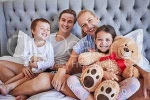 Portrait of a happy caucasian family with two children sitting on a bed holding teddybear and smiling at the camera. Loving parents spending free time with their daughter and son on the weekend
