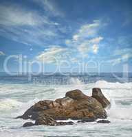 Copy space of a turbulent sea with rough tides and choppy waves from strong winds crashing onto big boulders at the beach with a cloudy blue sky background. Rocky coast in Western Cape, South Africa