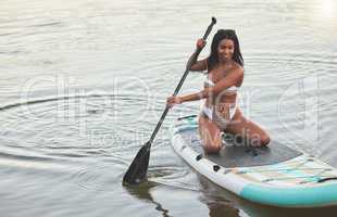 Young active african female smiling and kneeling on her paddle board on a lake out in nature. Young female enjoying a summer day at dawn on a paddle board. Life is better when youre paddling in the water