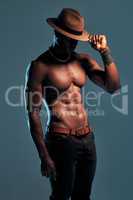 Fashionable african American model posing shirtless against blue studio background with copyspace. Sexy, serious black man with attitude showing bare six pack while wearing hat. Masculine and muscle