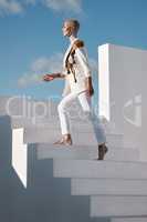 Escaping the ordinary. a young model wearing stylish white clothes while walking up stairs against a sky background.