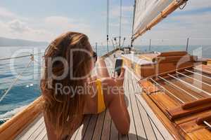 Young woman in yellow swimsuit sunbathing on boat deck using her cellphone to send text messages. Young woman using her cellphone while sunbathing in yellow swimsuit on boat.