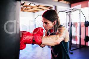 One fit young caucasian man wearing gloves and boxing a punching bag while training in a gym. Strong focused boxer practising his stance and technique for a competition fight. Fierce fighter striking punchbag with a hit and jab while learning self defence