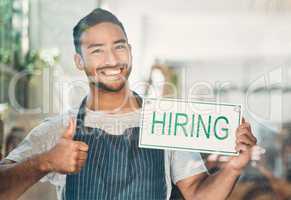 Portrait of one young hispanic man showing thumbs up while holding a hiring sign at a window on display in a cafe or store. Mixed race guy advertising job opportunity while recruiting new staff for his startup shop