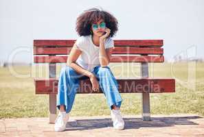 Cool young hispanic woman wearing sunglasses and sitting on a park bench outside. Carefree young woman with a curly afro wearing trendy, stylish sunglasses while enjoying a sunny day at the park