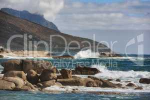 Ocean view of waves crashing against boulders or rocks in the sea in Camps Bay, Cape Town in South Africa. Relaxed and calm landscape view of the sea and mountains against a cloudy overcast sky