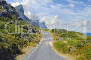Asphalt road on Table Mountain with cloudy blue sky. Landscape of countryside roadway for traveling on mountain pass along a beautiful scenic nature drive with lush shrubs in Cape Town, South Africa