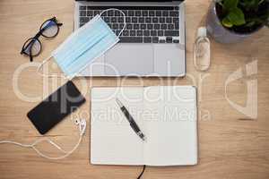 Office desk layout of laptop keyboard, face mask, hand sanitiser, glasses, smartphone with earphones and notebook with pen during the covid-19 pandemic