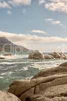 Rocks in the ocean under a blue cloudy sky with copy space. Scenic landscape of beach waves splashing against boulders or big stones in the sea at a popular summer location in Cape Town, South Africa