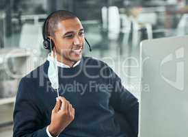 Young male call center agent smiling and wearing a headset and mask working on a computer in an office at work. Customer service, support and sales. Giving advice and helping. Answering calls