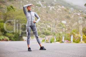 Active and fit young woman taking a break from exercising outdoors. Athletic female wiping sweat from her forehead after a jog or run outside. Woman looking tired but determined to continue her workout