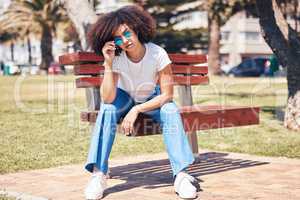 Young stylish mixed race woman with curly afro hair wearing trendy sunglasses and relaxing on a bench at the park. One female only looking carefree, cool and confident while enjoying sunny day outside