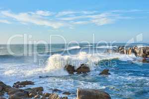 A rocky coastline in the Cape Province, South Africa. Ocean waves crashing on coastal rocks on a sunny summer day with blue clear skies and a scenic tropical landscape beachfront in the Western Cape