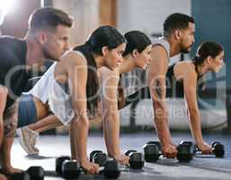 Diverse group of active young people doing plank hold and push up exercises with dumbbells while training together in a gym. Focused athletes doing press ups and renegade rows with weights to build muscle and endurance during a workout in a fitness class