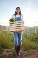 Portrait of a woman farmer holding a wooden box of fresh vegetables. Young brunette female standing on a field while holding a basket of organic produce