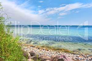 Copyspace at sea with a cloudy blue sky background. Calm ocean waves washing onto stones at an empty beach shore with sailboats cruising in the horizon. Scenic landscape for a relaxing summer holiday