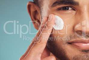 Closeup of one young indian man applying moisturiser lotion to his face while grooming against a blue studio background. Handsome guy using sunscreen with spf for uv protection. Rubbing facial cream on cheek for healthy complexion and clear skin