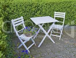 Two courtyard chairs and table in a secluded and private garden at home. Wooden patio furniture and seating area for coffee and reading in a backyard. Purple meadow cranesbill flowers and a hedge