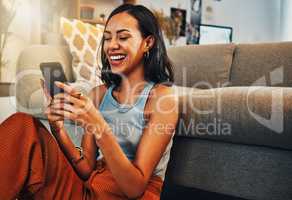 Beautiful mixed race woman browsing internet on cellphone in home living room. Happy hispanic sitting alone on floor in lounge and using technology to network. Laughing while scrolling on social media