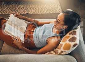 Above view of mixed race woman reading book in a living room at home. Beautiful hispanic sitting alone on lounge sofa and enjoying novel. Relaxed woman wearing glasses to read fiction story on weekend
