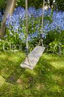 Fun garden swing for children to relax, enjoy and play on a weekend or break with common bluebell flowers growing in background. Homemade wood and rope playset in a home backyard or park playground