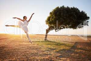Passionate young female ballerina dancing outside in nature environment with a tree in the background. Ballet dancer jumping in front of a bended tree on a sunny day