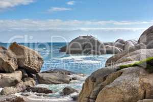 Beautiful sea view of big boulders and ocean water on a sunny beach day in summer. A seaside view of nature with a blue sky, white clouds, and waves. A seascape near the shore under the horizon