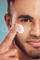 Closeup of one young indian man applying moisturiser lotion to his face while grooming against a blue studio background. Handsome guy using sunscreen with spf for uv protection. Rubbing facial cream on cheek for healthy complexion and clear skin