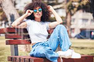 Young stylish mixed race woman with curly afro hair wearing trendy sunglasses and relaxing on a bench at the park. One female only looking carefree, cool and confident while enjoying sunny day outside