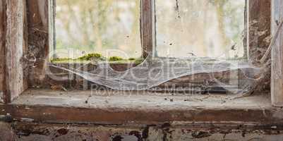 Abandoned, dirty and dusty window covered in spiderwebs in empty house from poverty and economic crisis. Old, damaged and weathered wooden windowsill and wood frame rotting from dampness and neglect
