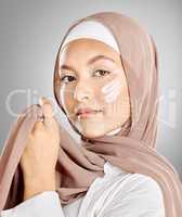 Portrait of a Muslim woman using face moisturiser, sunscreen or lotion isolated on a grey studio background with copyspace. Young female wearing a hijab or headscarf while doing her skincare routine