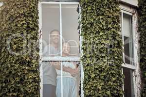 Happy young mixed race couple standing in their window. Loving couple bonding and spending time together at home. Lovers standing by a window with leaves growing up the exterior wall. Warm and cozy at home