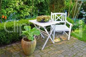 Patio chair and square table with lots of plants in a garden. Outdoor furniture for relaxing in summer while flower gardening. Potted plants in a small paved courtyard with green grass and trees