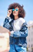 Trendy and cool hispanic woman wearing sunglasses and denim casual clothes while standing outside. Cheerful woman with a curly afro looking fashionable while enjoying a sunny day outside