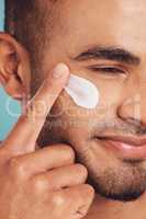 Closeup of one young indian man applying moisturiser lotion to his face while grooming against a blue studio background. Handsome guy using sunscreen with spf for uv protection. Rubbing facial cream on cheek for healthy complexion and clear skin